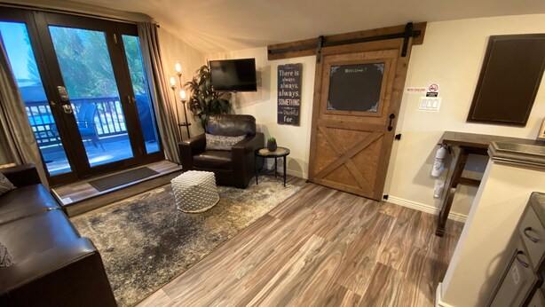 A cozy living room with a leather sofa, a round ottoman, a television mounted on the wall, and a sliding barn-style door. - Cabernet Cottage (Cottonwood)