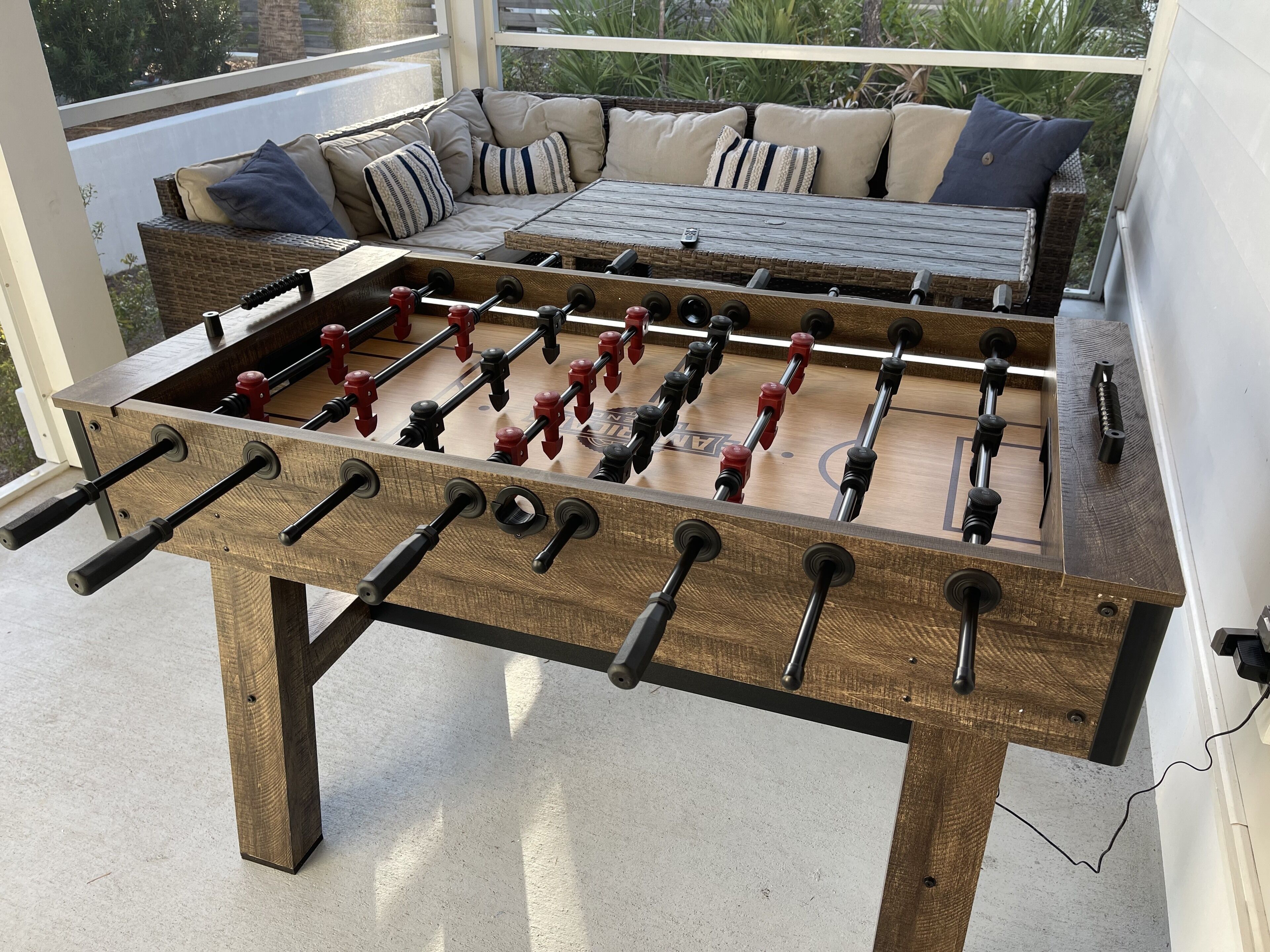 Une table de baby-foot dans une pièce avec un canapé et des coussins, une fenêtre donnant sur un paysage verdoyant et un sol en béton.
