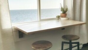 Een kamer met een groot raam met uitzicht op zee, een houten bureau, twee krukken en een plant in een pot.