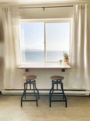 A room with a window overlooking the sea, two stools, and a small plant on the windowsill.