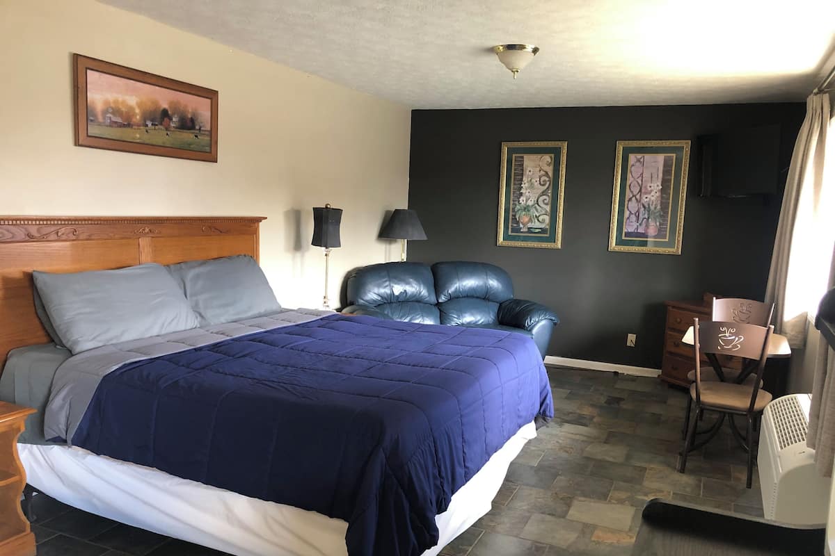 A hotel room with a bed, a blue leather chair, a wooden nightstand, a desk with a lamp, and two framed pictures on the wall.