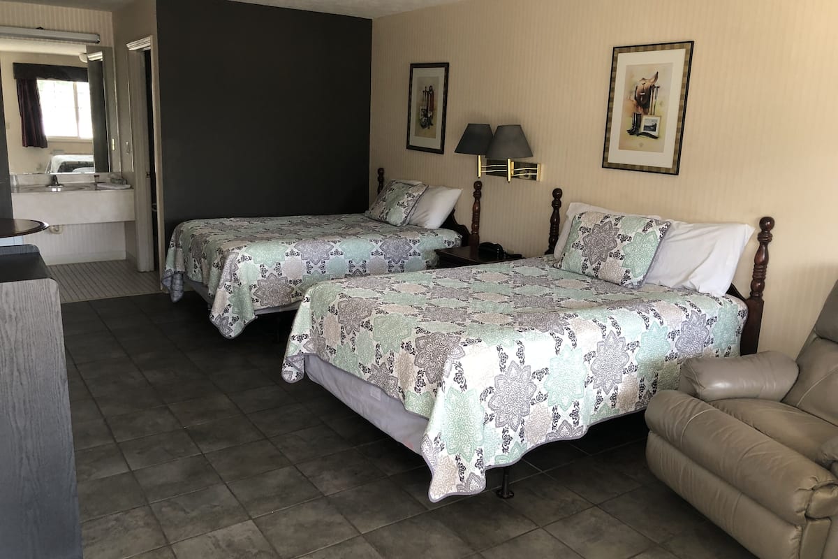 A hotel room with two beds, a sofa, a desk with a lamp, and a bathroom visible through an open door.