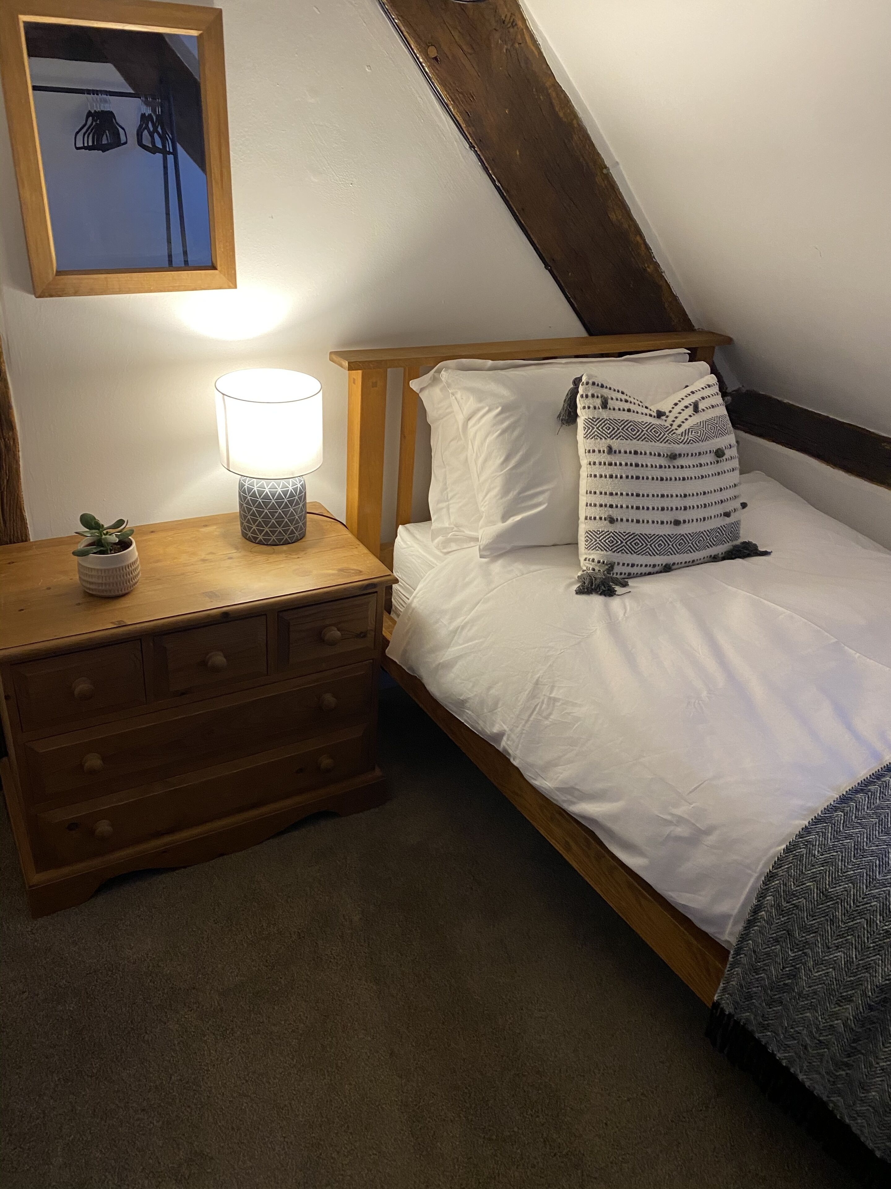 A bedroom with a wooden bed, white bedding, a bedside lamp, a small potted plant, and a wooden dresser.
