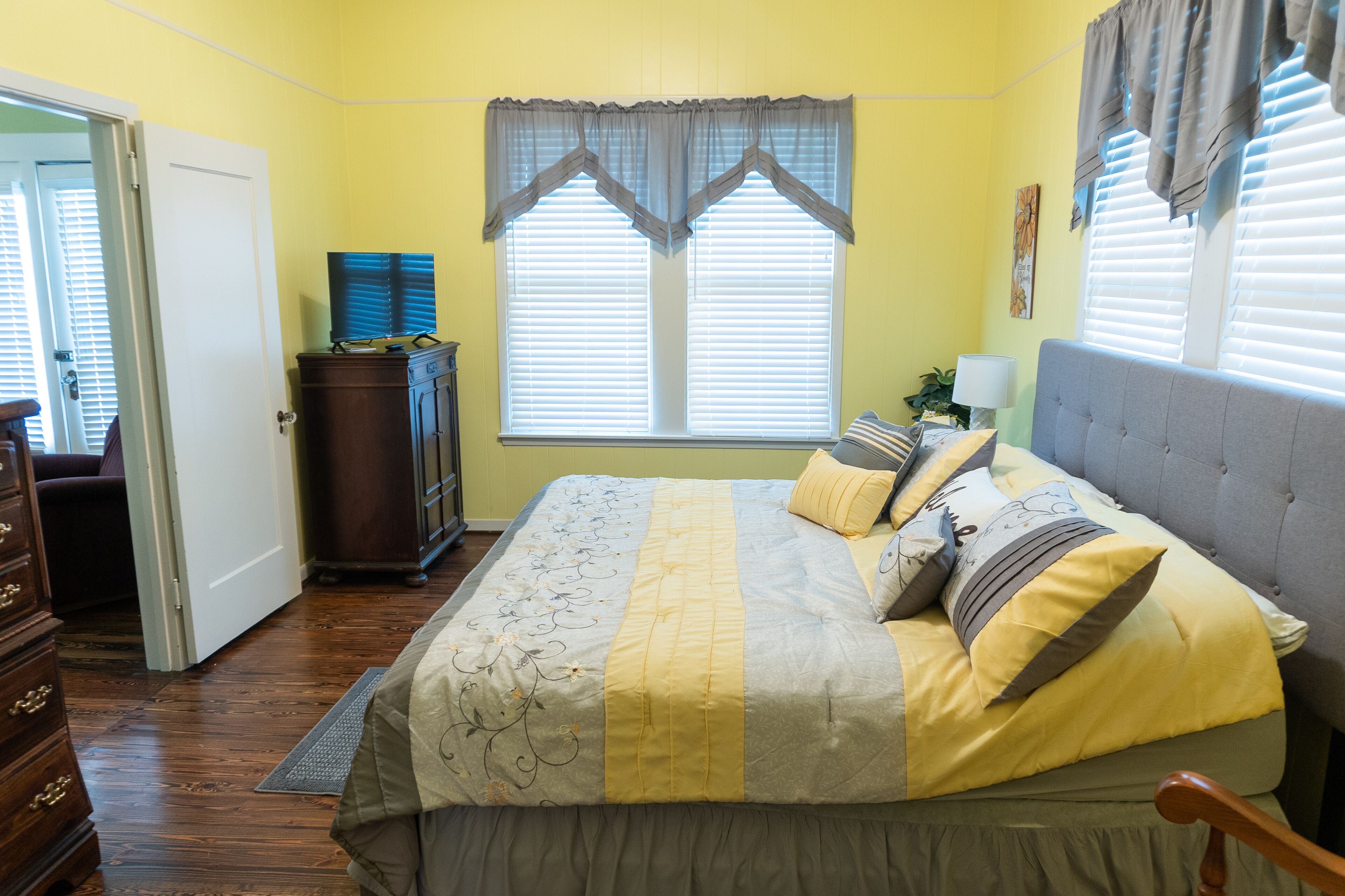 A bedroom with a bed, a dresser, a window with blinds, and a door leading to another room.