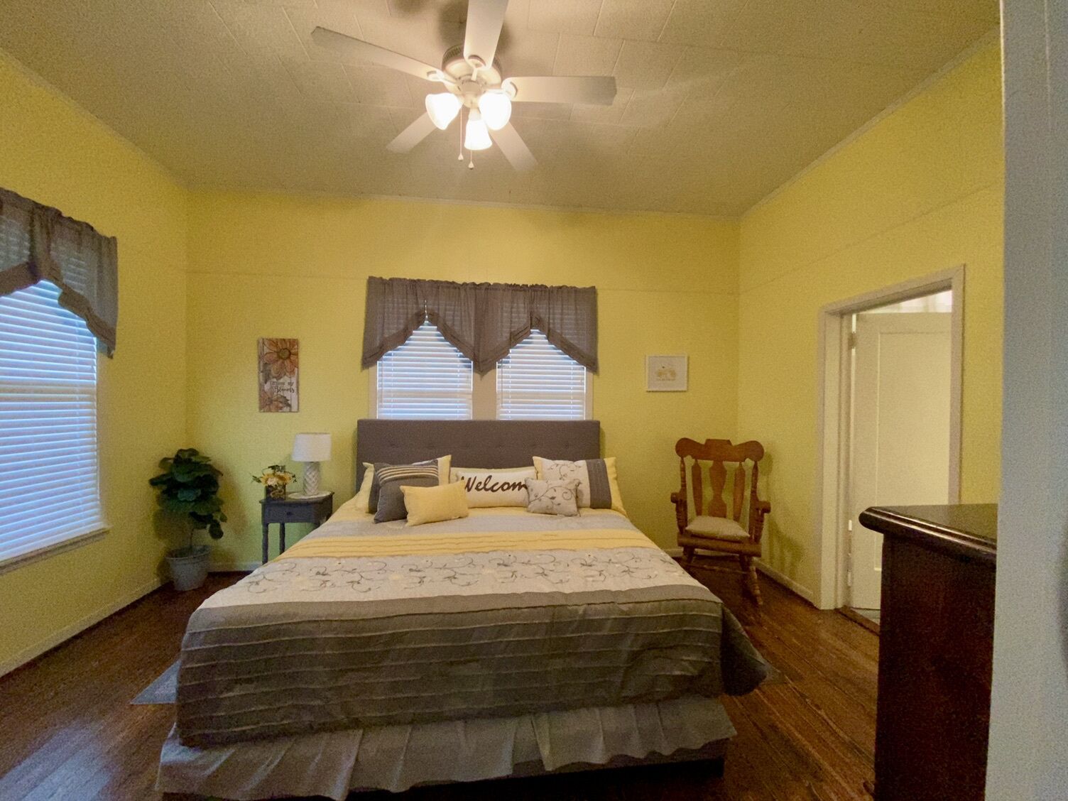 Un dormitorio con cama, ventilador de techo, ventana con cortinas, una silla y una puerta.
