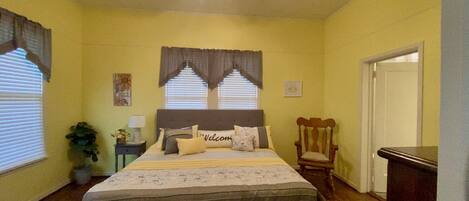 Un dormitorio con cama, ventilador de techo, ventana con cortinas, una silla y una puerta.