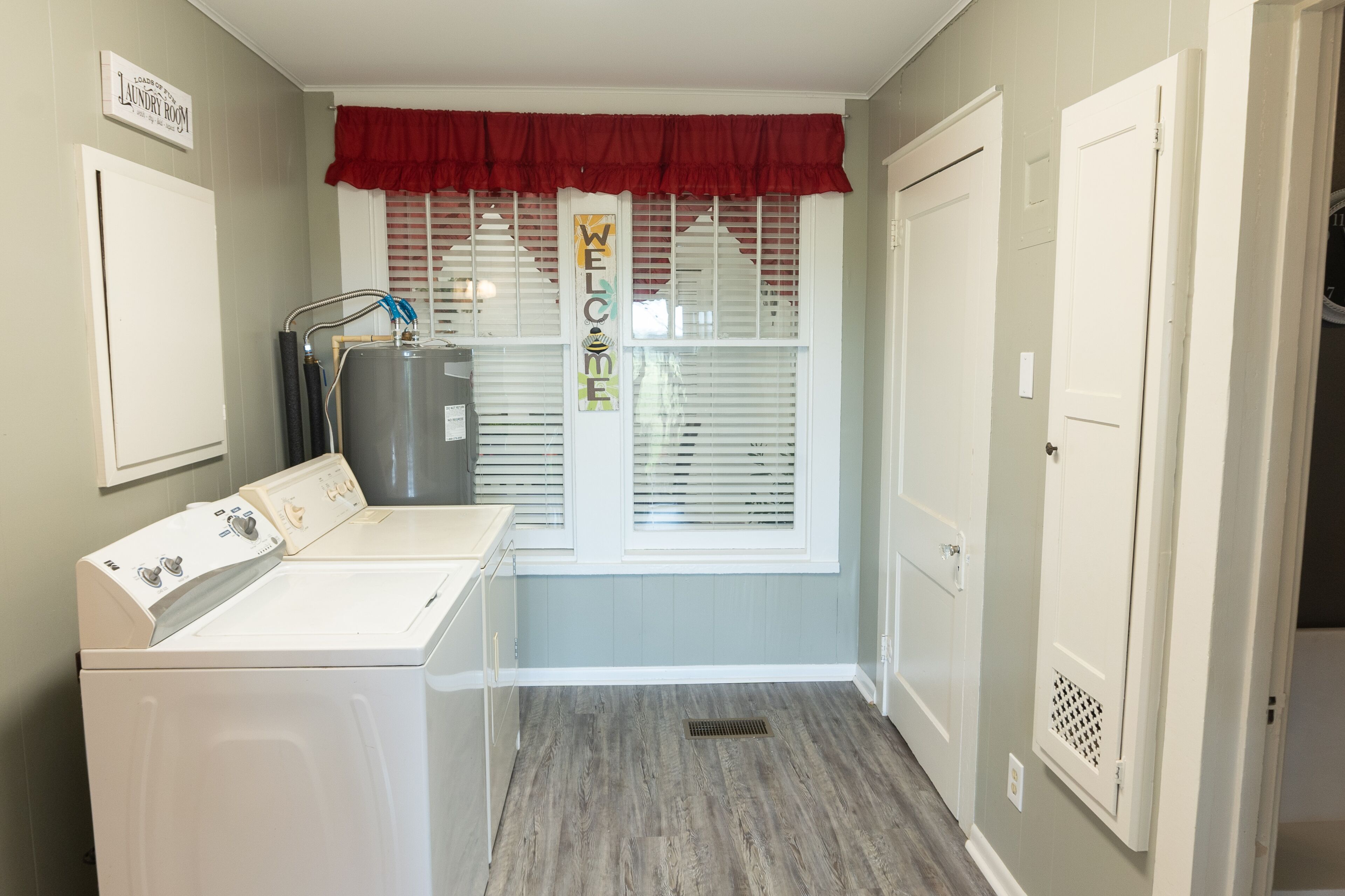 A laundry room with a washing machine, dryer, and water heater. There is a window with blinds and a red valance.