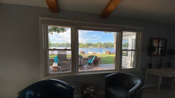 A room with a large window overlooking a lake, two black chairs, a small table, and a wooden ceiling beam.