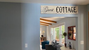 A room with a 'Guest Cottage' sign, a white table with a bottle, a fan, and a chair with colorful stripes.