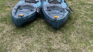Two kayaks on a grassy lawn with a house and a tree in the background.