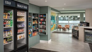 A hotel lobby with a cold beverage refrigerator, snack shelves, a mural, and seating area.