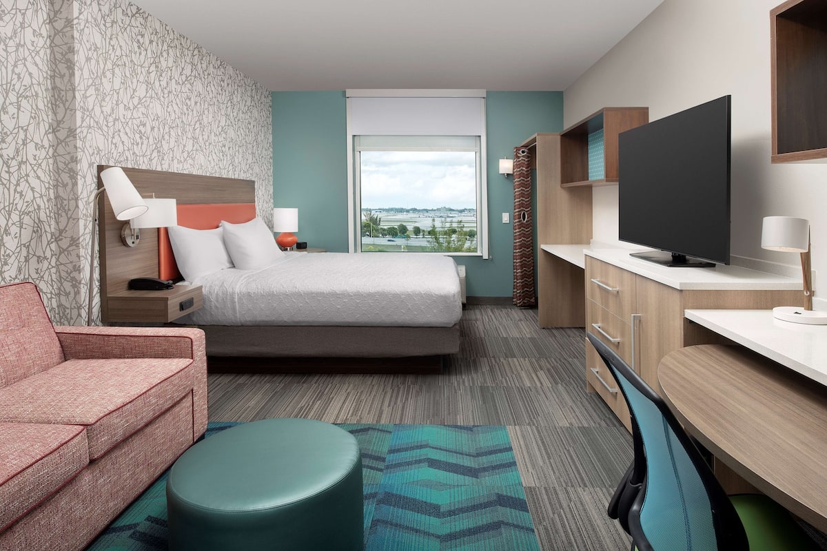 A hotel room with a bed, a sofa, a desk, a TV, and a window with a city view.