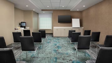 Una sala de conferencias con alfombra estampada, sillas negras, una estación de café, una pizarra y un televisor de pantalla plana en la pared.