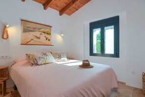 Un dormitorio con cama, ventana, un cuadro y un sombrero sobre la cama.