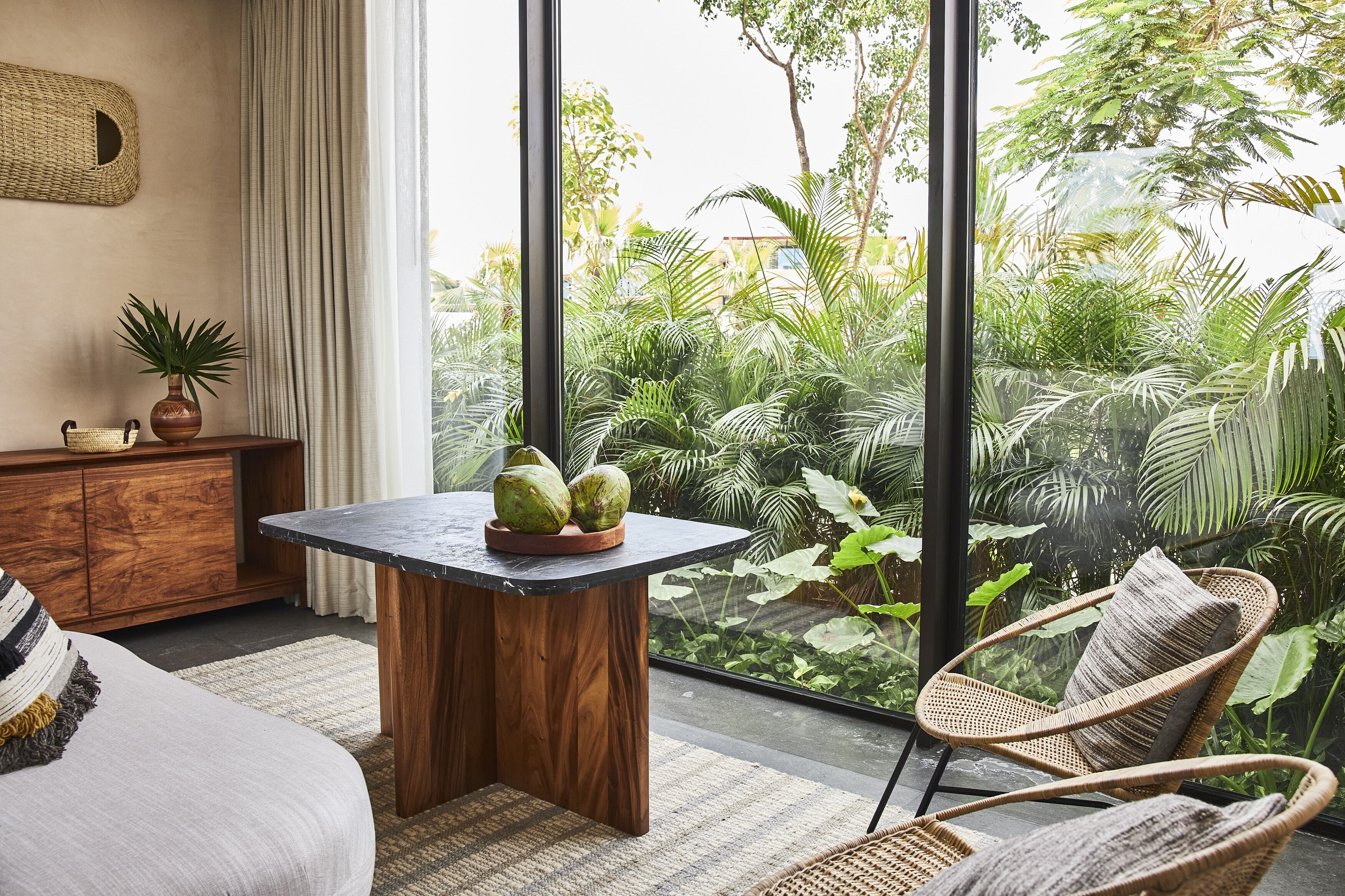 A room with a large window, a wooden table with a vase and coconuts, a wicker chair, and a sofa with a striped pillow.