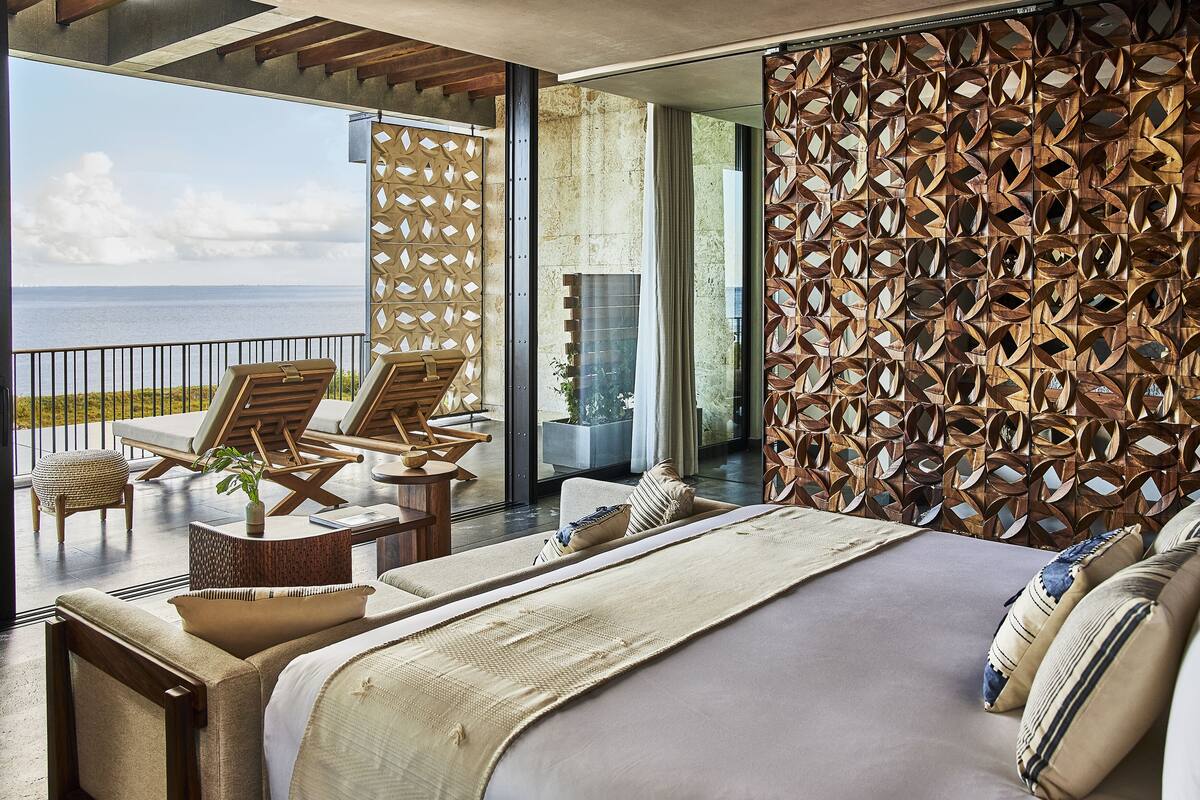 A modern bedroom with a large bed, a balcony with lounge chairs, and a view of the sea.