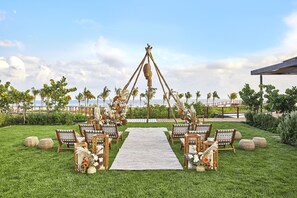 An outdoor wedding setup with a white aisle carpet, wooden chairs, and a decorative arch.