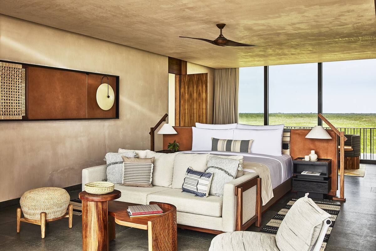 A modern hotel room with a large bed, a sofa, a coffee table, and a view of the outdoors.