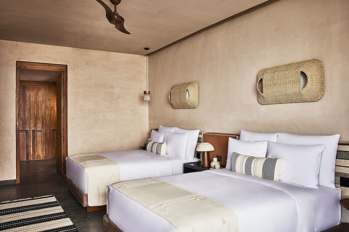 A hotel room with two beds, a wooden door, a woven wall decoration, and a nightstand with a lamp.