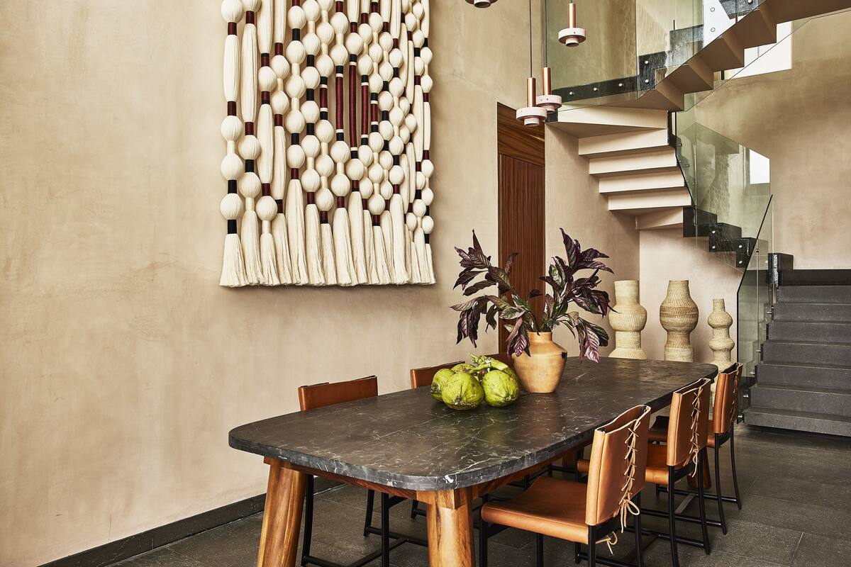 A dining area with a marble table, wooden chairs, a decorative wall piece, and a staircase.