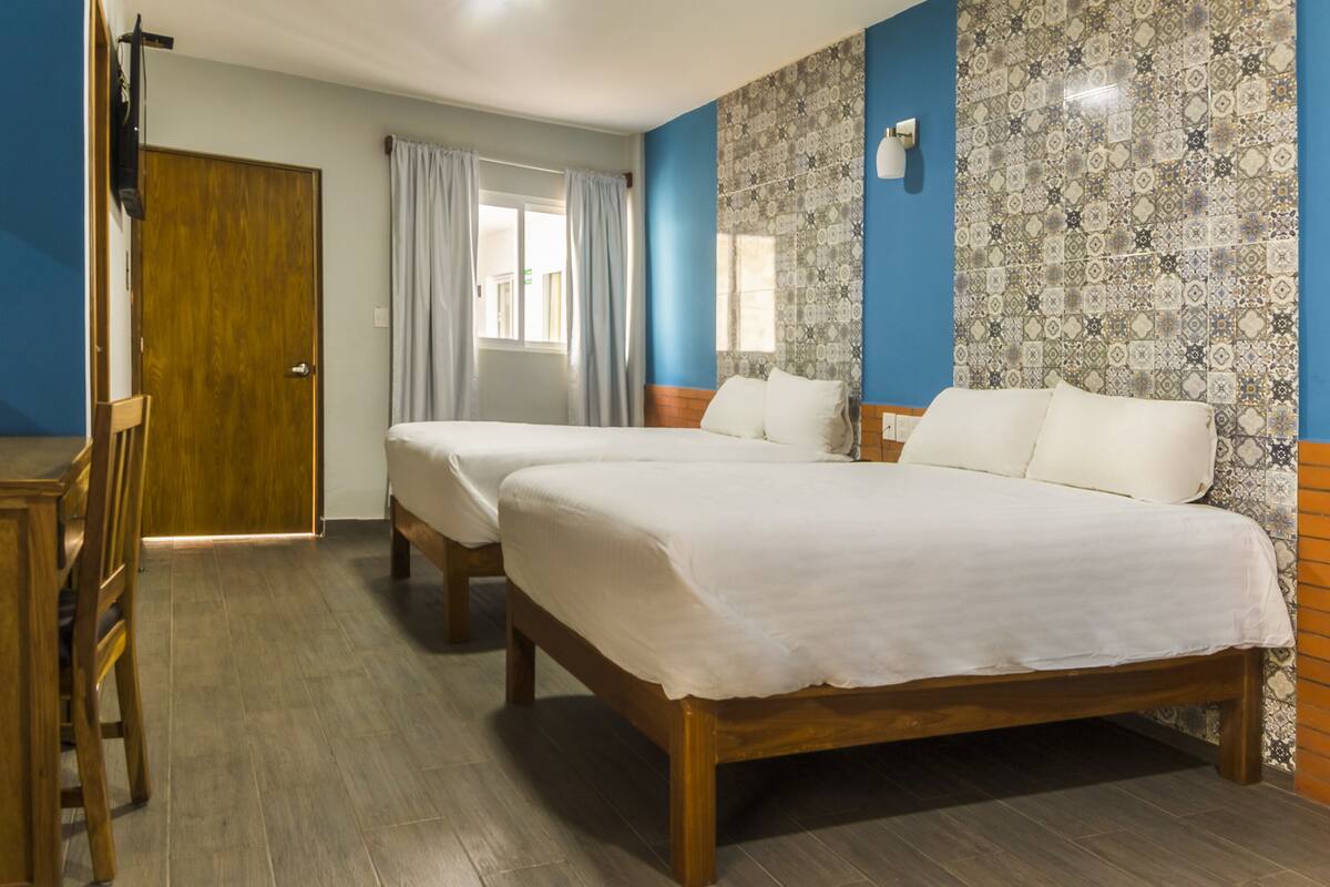 A hotel room with two single beds, a wooden desk, a chair, a window with curtains, and patterned tile walls.