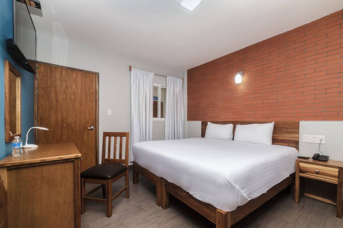 A hotel room with a wooden bed, a wooden nightstand, a wooden chair, a wooden desk, a mirror, and a window with white curtains.