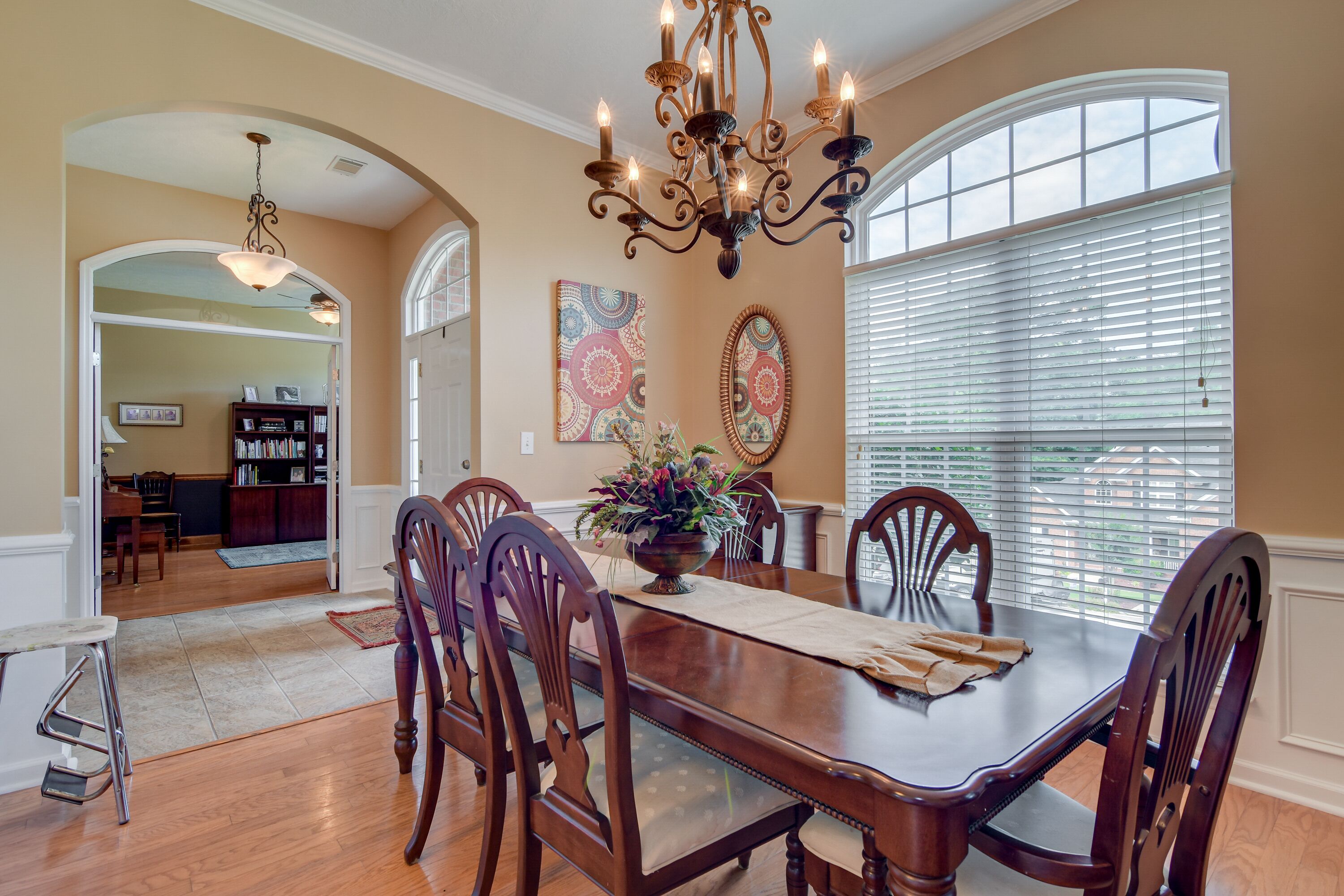 A dining area with a wooden table, chairs, and a chandelier. There are two large windows with blinds, a rug on the floor, and a door leading to another room.