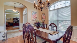 A dining area with a wooden table, chairs, and a chandelier. There are two large windows with blinds, a rug on the floor, and a door leading to another room. - Master's Rental (Evans)