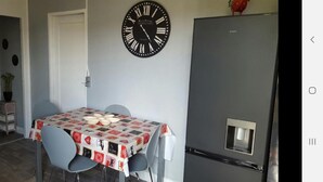 A dining area with a table covered by a patterned tablecloth, two chairs, a wall clock, and a refrigerator. - Charming little house "LA BIKOK (BILLY)