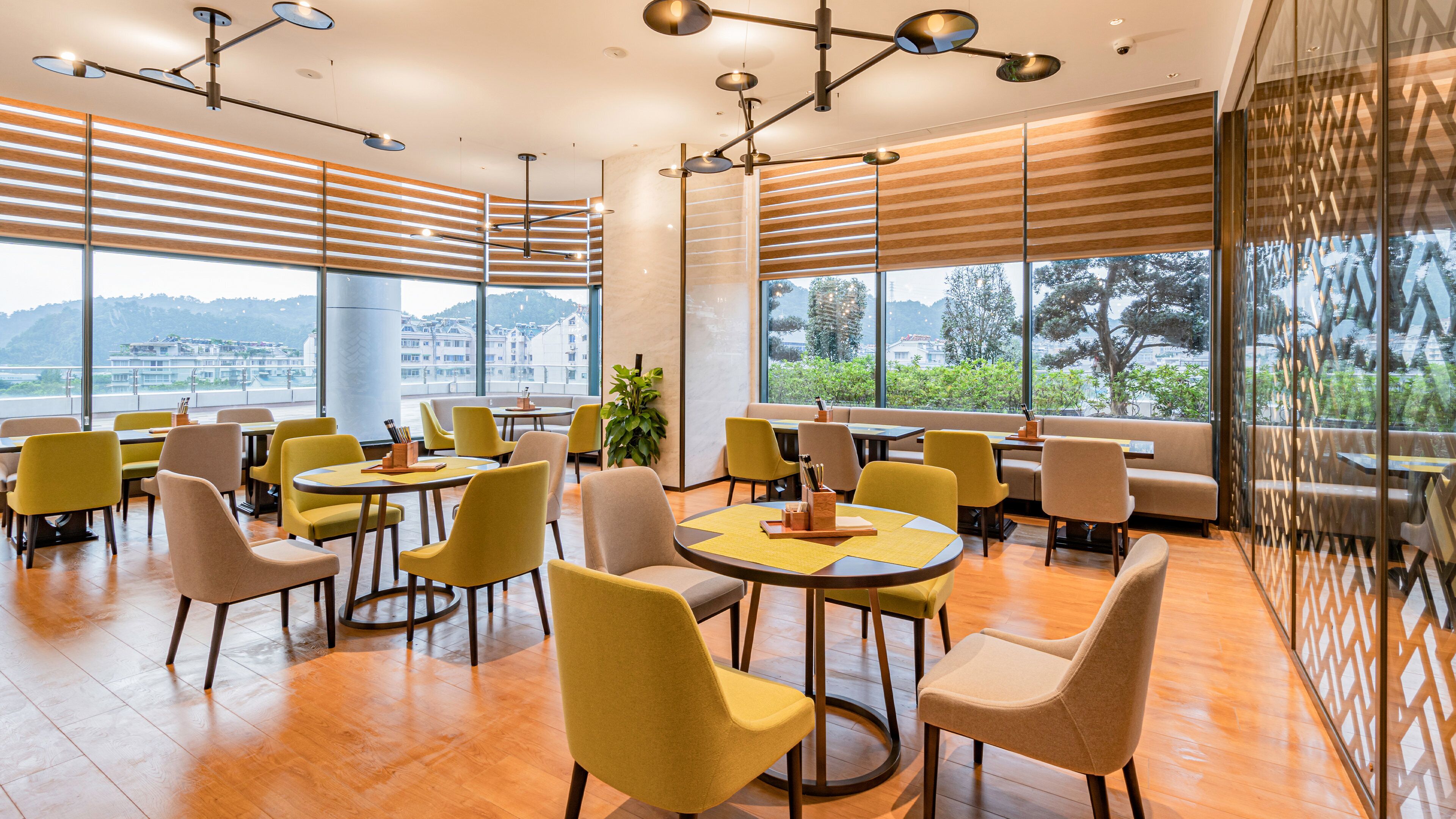 A modern dining area with round tables and chairs, large windows with a view of buildings and trees, and a ceiling with pendant lighting.