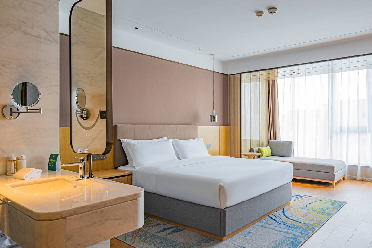 A modern hotel room with a large bed, a sitting area, a bathroom with a vanity, and a large window with curtains.