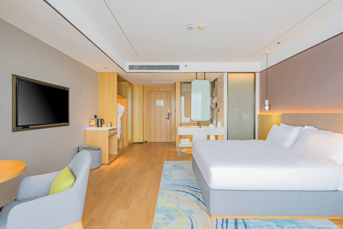 A modern hotel room with a large bed, a flat-screen TV, a desk with a chair, and a bathroom with a mirror and sink.