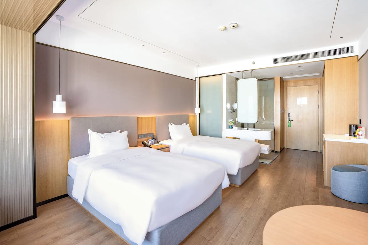 A modern hotel room with two beds, a desk, a mirror, and a small seating area.