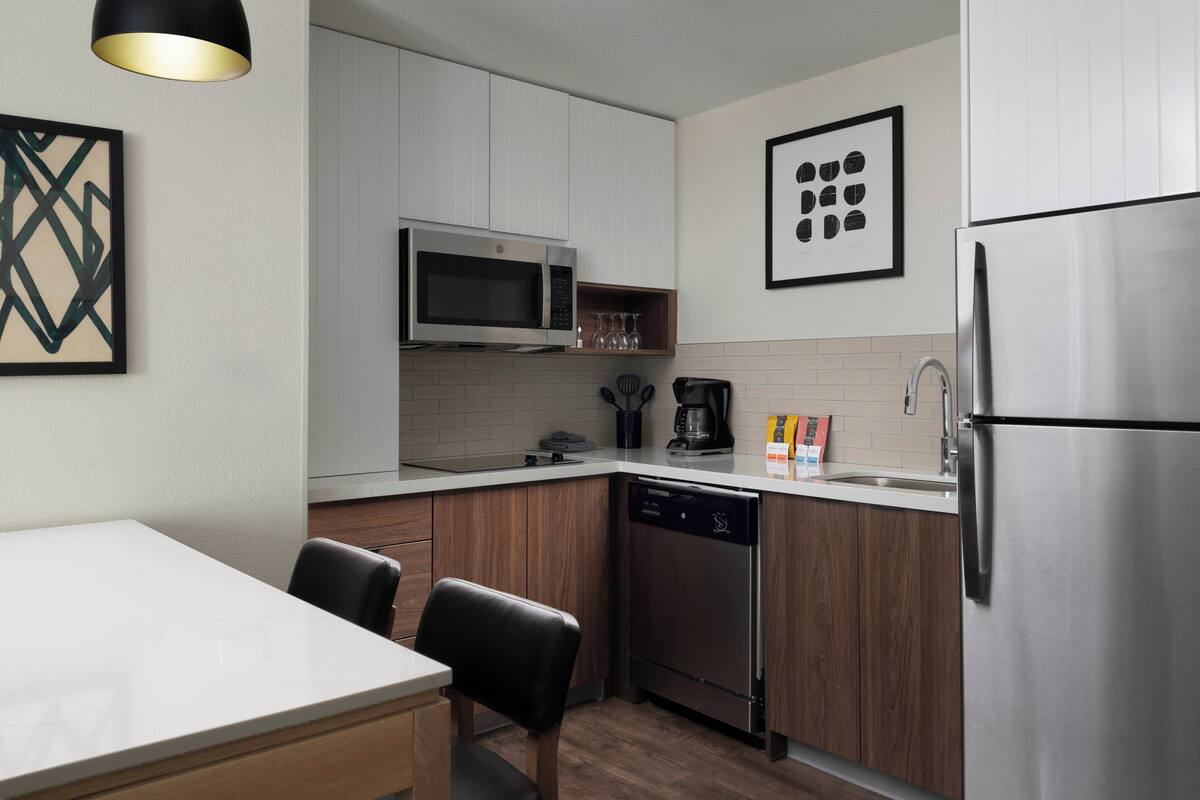 A compact kitchen with white cabinets, a stainless steel refrigerator, a microwave, and a dishwasher. There are two black chairs at a white table, and two pendant lights above the counter.