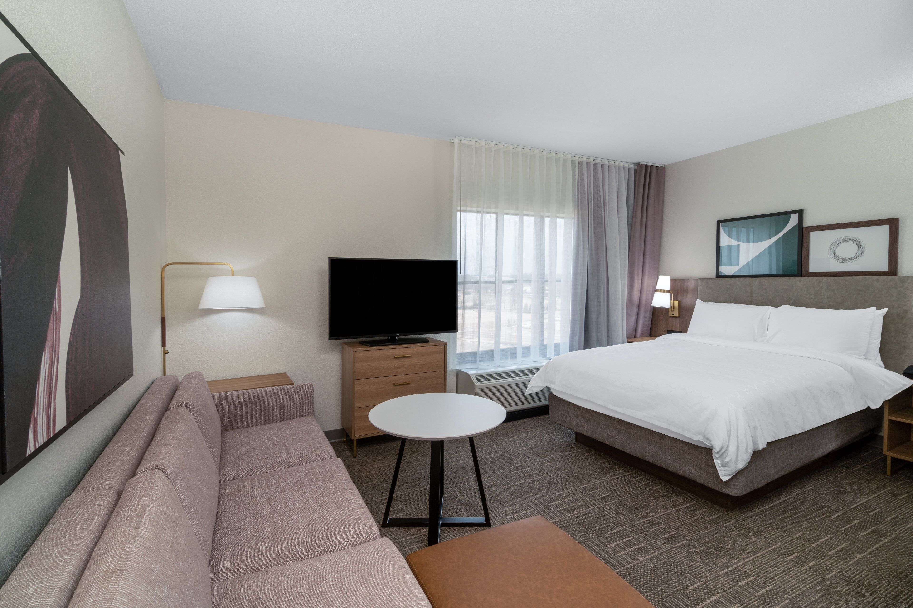 A hotel room with a bed, a sofa, a TV, a small table, and a window with curtains.