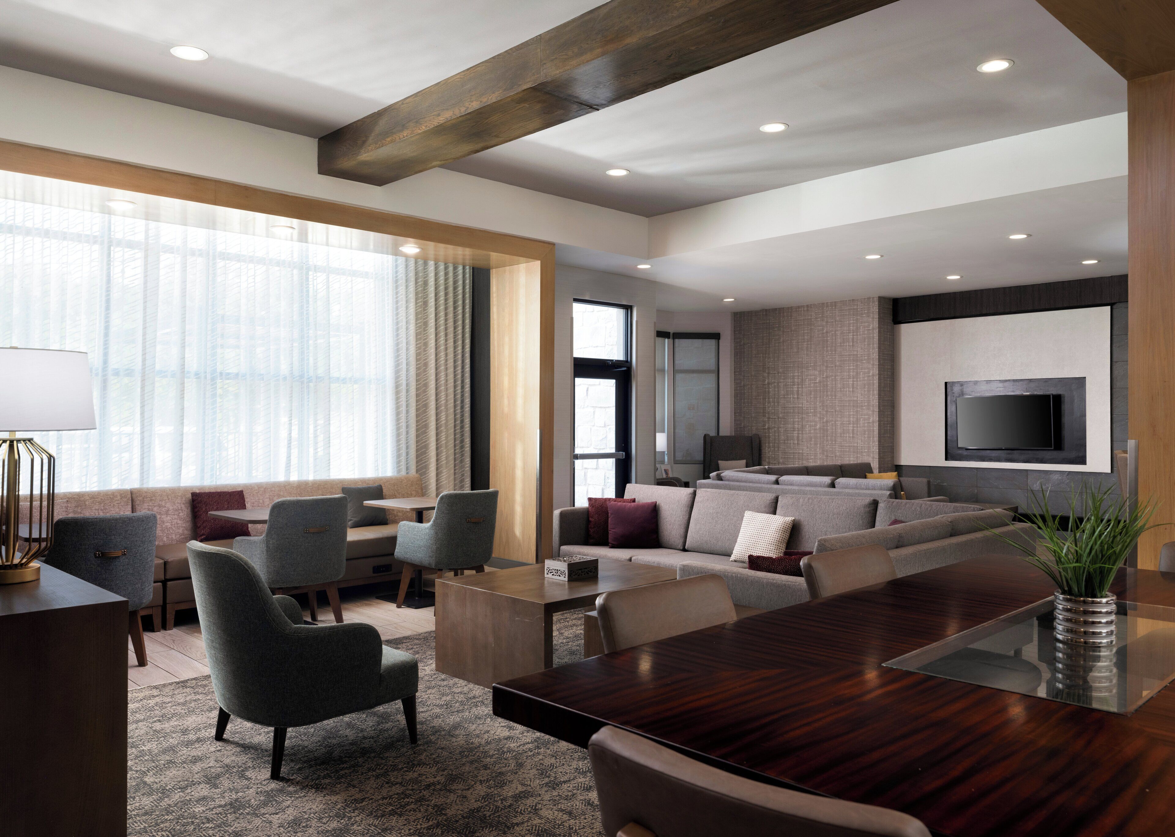 A modern living room with a large wooden table, comfortable seating, a flat-screen TV mounted on the wall, and large windows with sheer curtains.