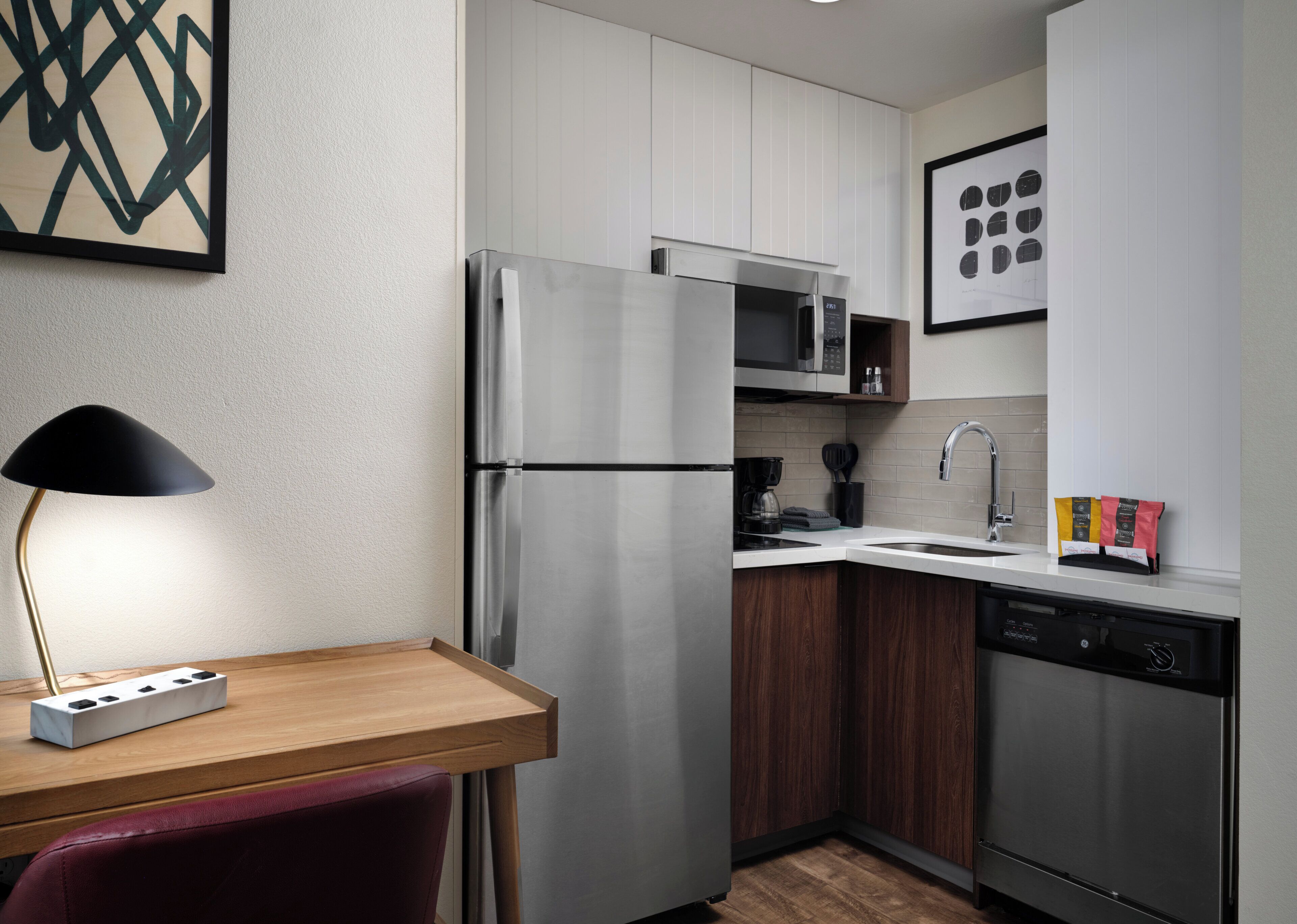 A modern kitchen with a stainless steel refrigerator, microwave, and dishwasher. There is a wooden table with a lamp and a power strip.