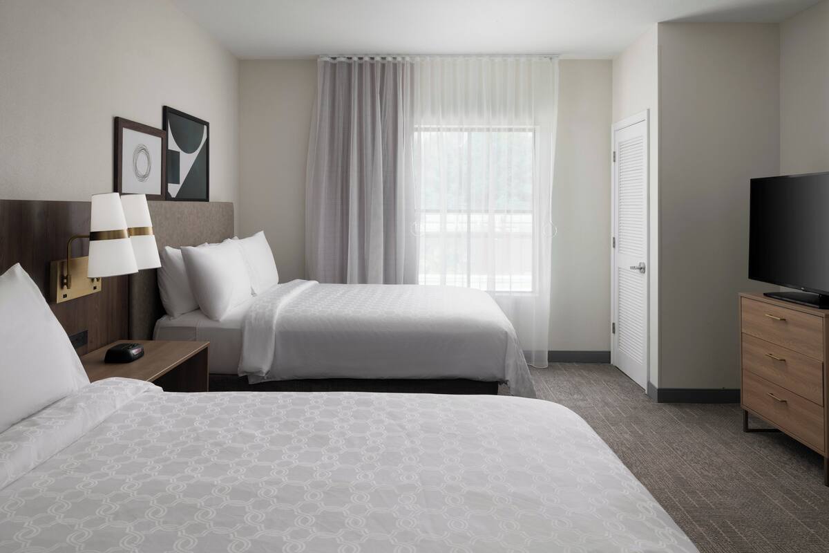 A hotel room with two beds, a TV, a desk with a lamp, and a window with curtains.