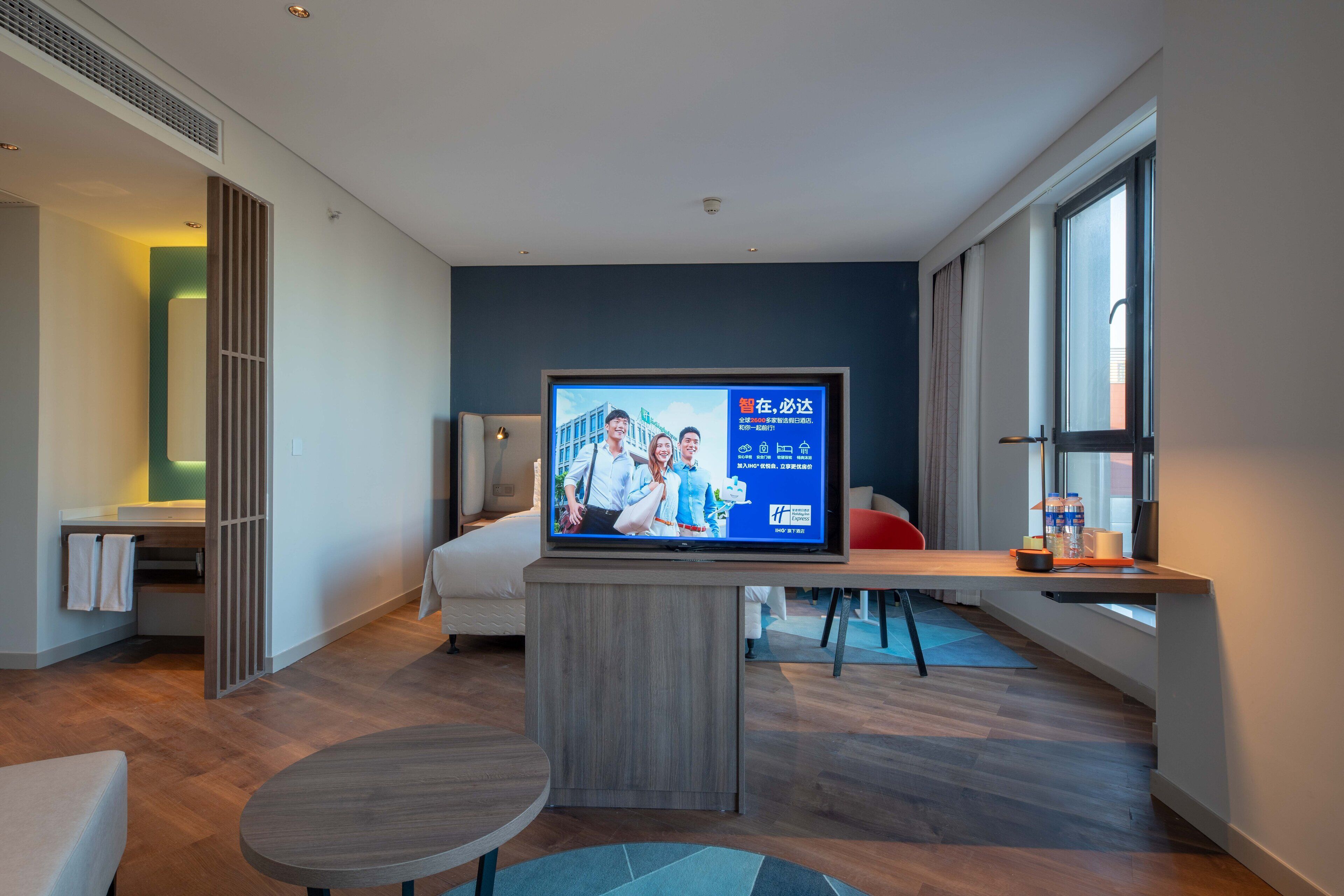 A modern hotel room with a large flat-screen TV, a bed, a desk, and a seating area.