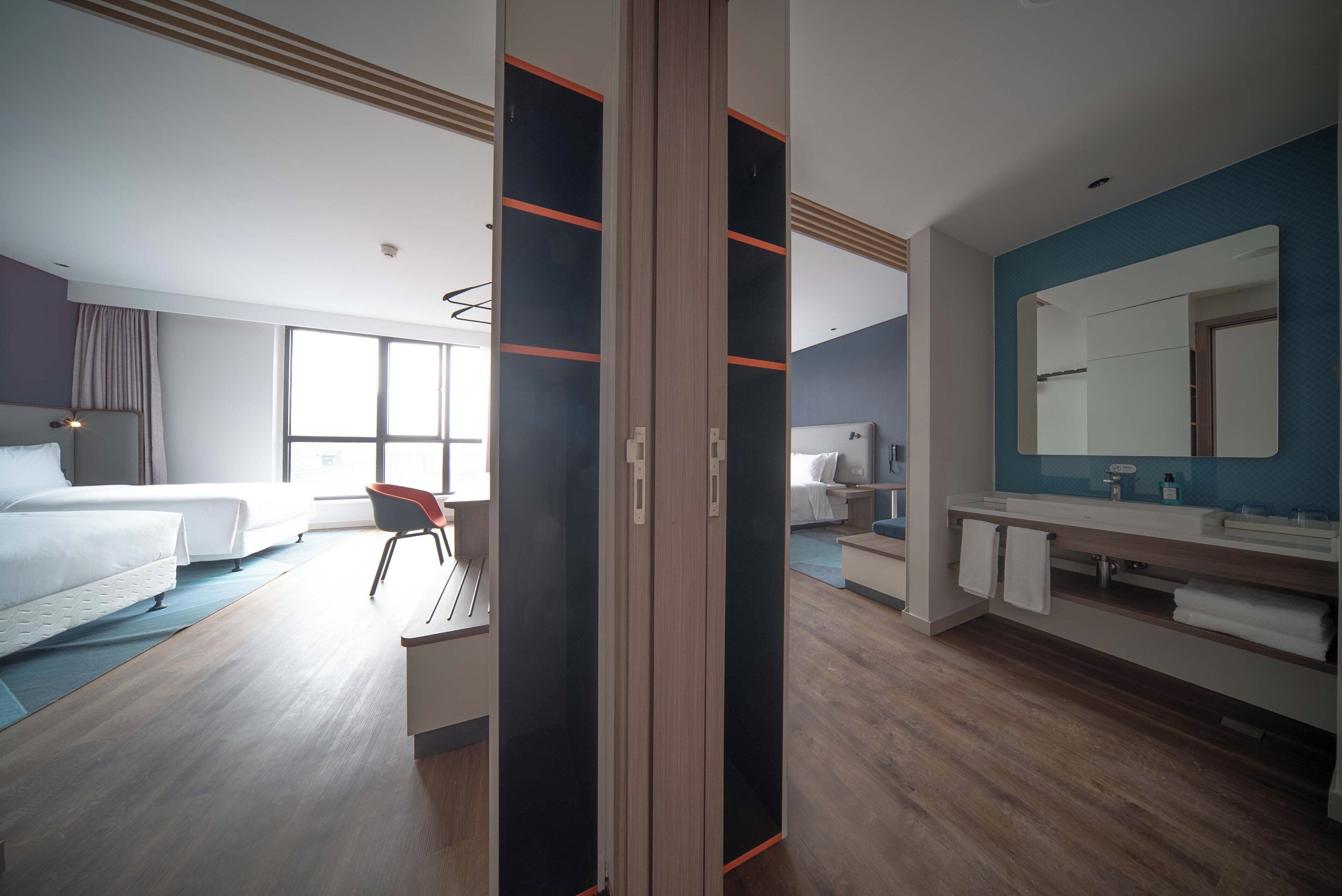 Une chambre d’hôtel moderne équipée d’un lit, d’un bureau, d’une chaise, d’une salle de bain avec un miroir et d’une grande fenêtre.