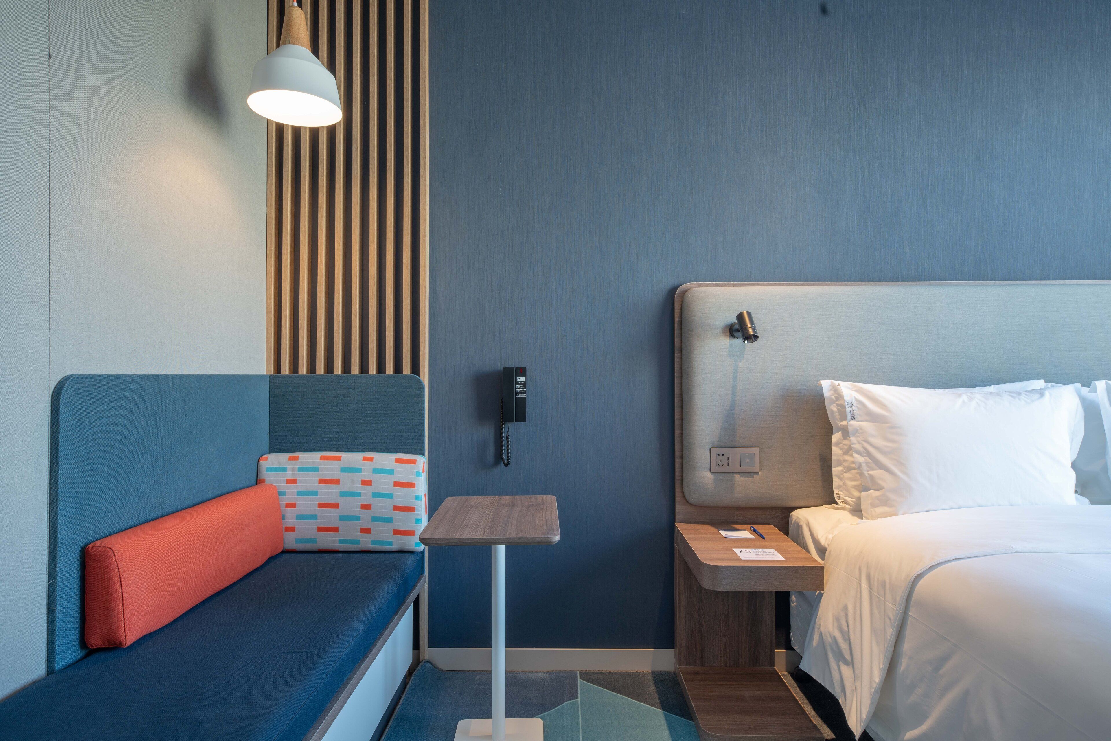 A hotel room with a bed, a small table, a nightstand, a wall-mounted phone, and a seating area with a cushion and a pillow.