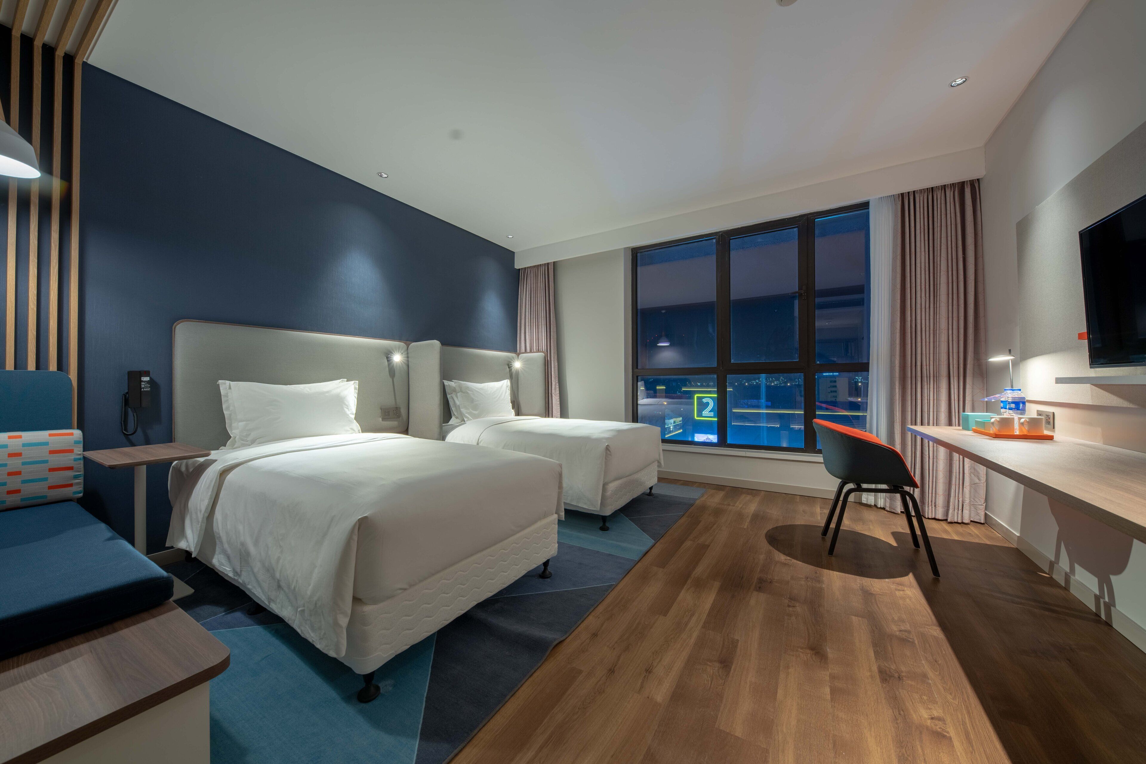 A hotel room with two beds, a desk, a chair, a TV, and a large window with a view of the city.