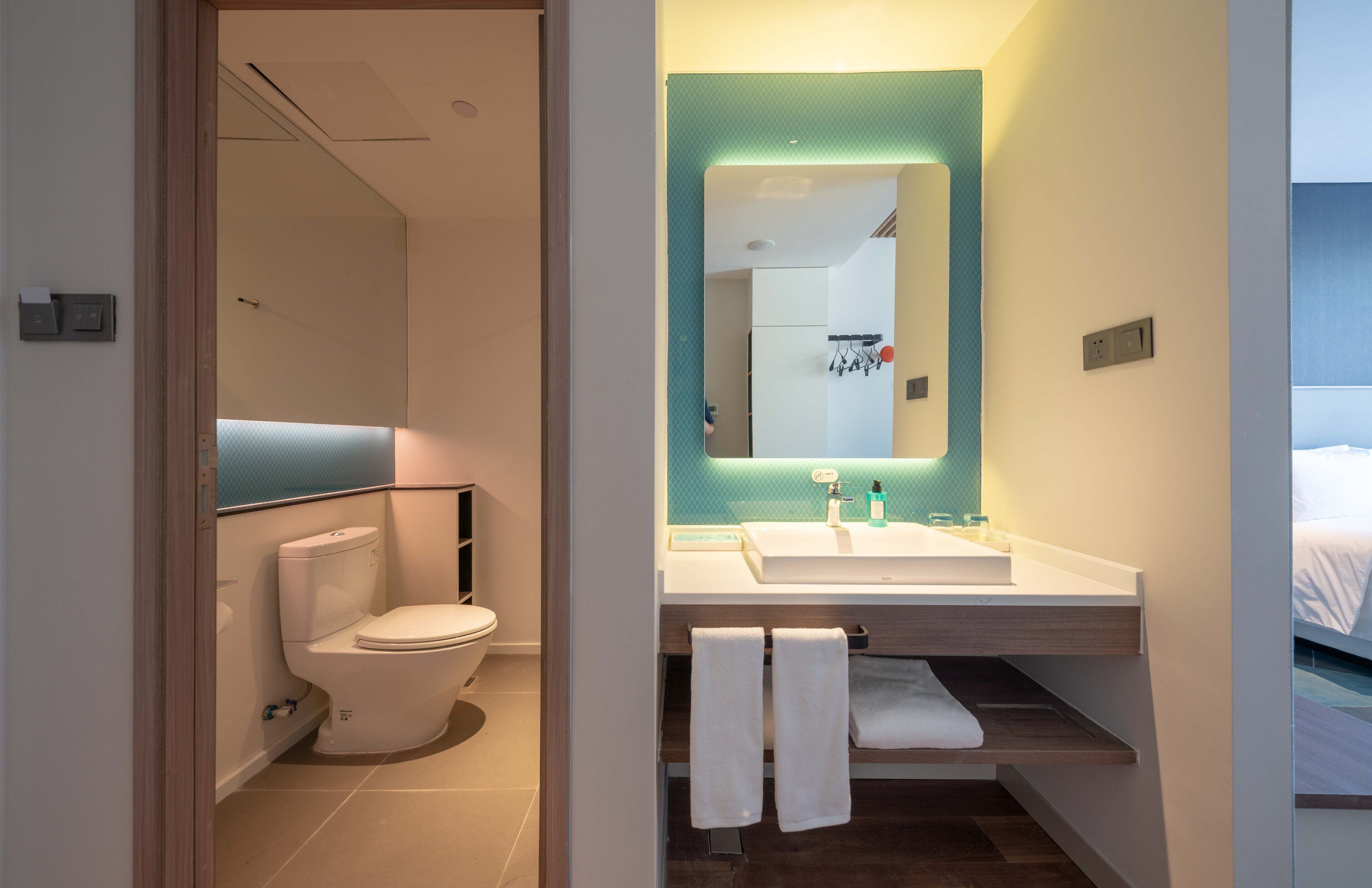 A modern bathroom with a toilet, sink, and mirror. There are towels hanging on a rack and a shelf with toiletries.
