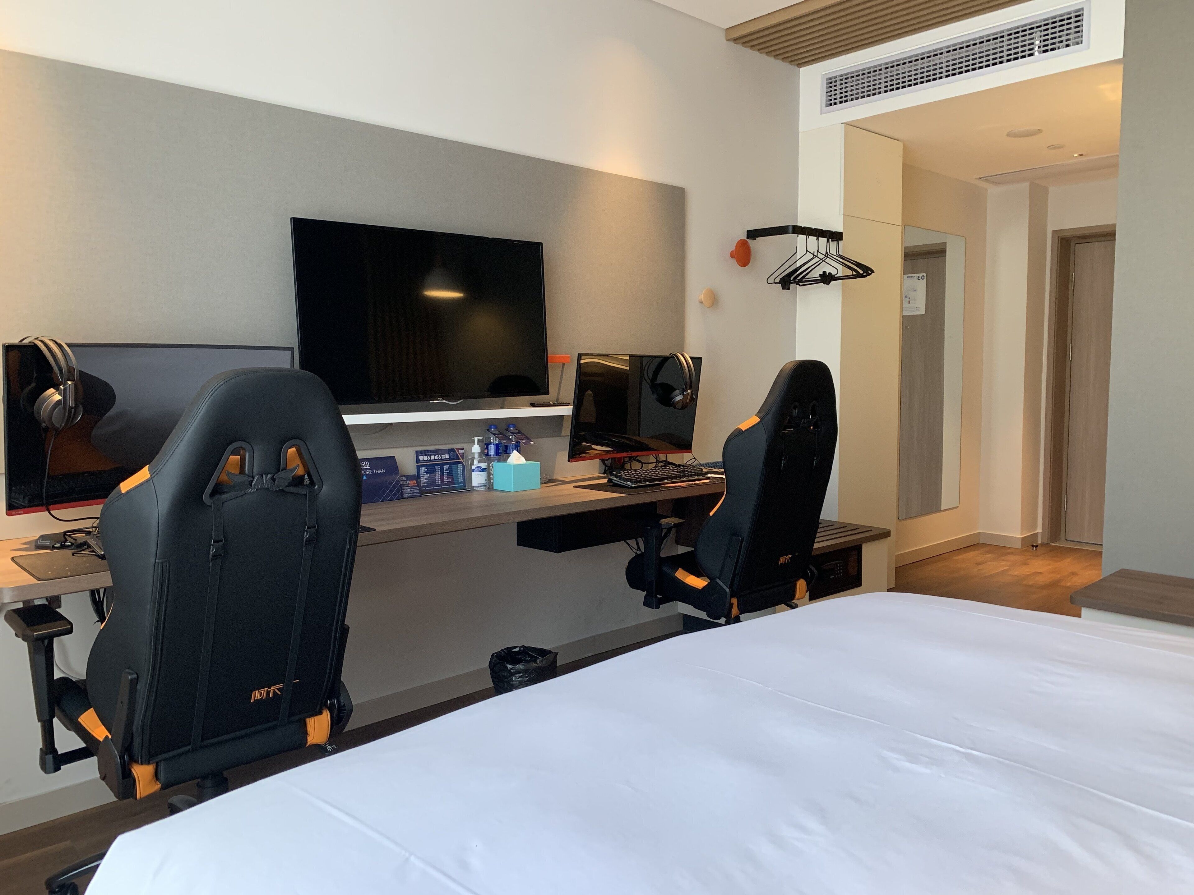 A hotel room with a large bed, two gaming chairs, a desk with a computer and headphones, a flat-screen TV, and a small table with bottles.