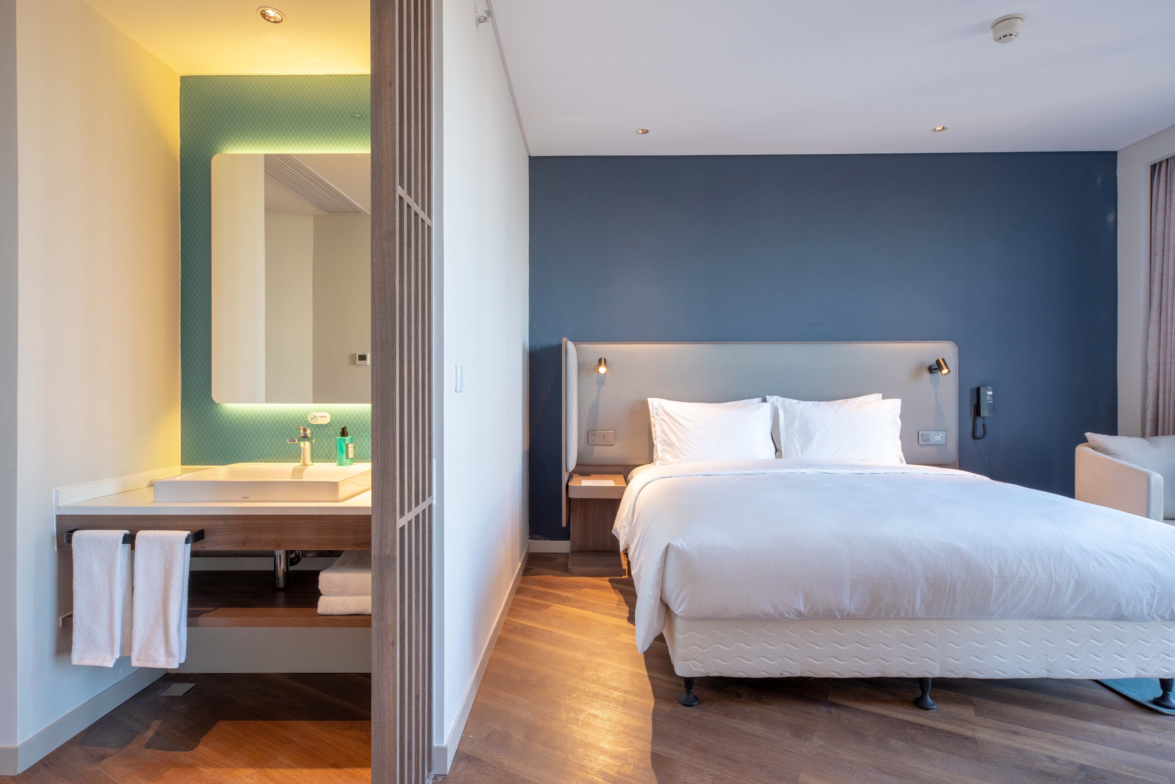 Quarto de hotel moderno com uma cama grande, uma mesinha de cabeceira, um armário para guardar objetos, banheiro com pia e espelho e piso de madeira.