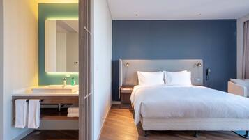 Quarto de hotel moderno com uma cama grande, uma mesinha de cabeceira, um armário para guardar objetos, banheiro com pia e espelho e piso de madeira.