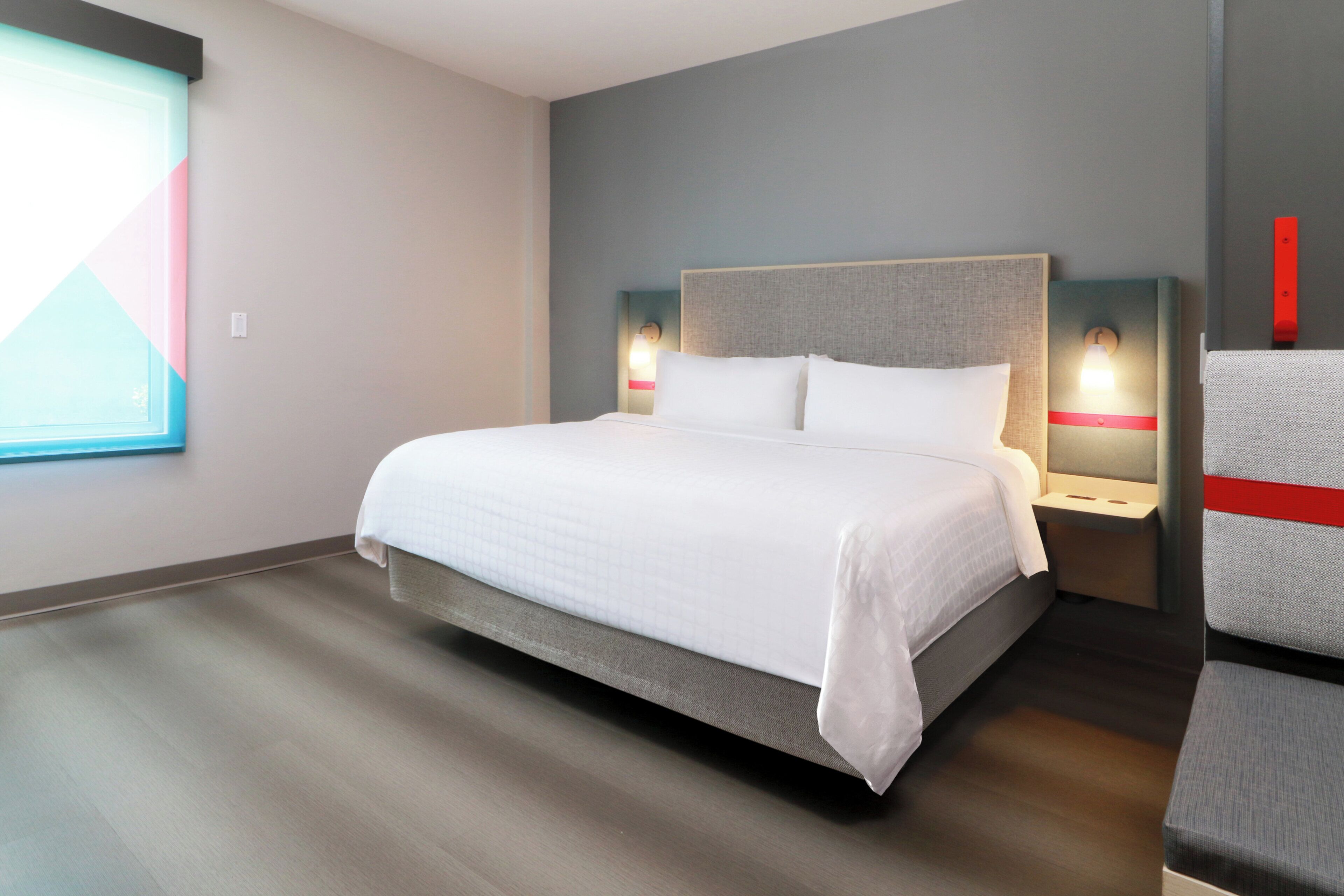 Una habitación de hotel moderna con una cama grande, dos mesitas de noche con lámparas, una ventana con persianas y vistas al exterior.