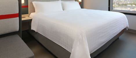 A hotel room with a bed, a nightstand, a chair, and a window with a city view.