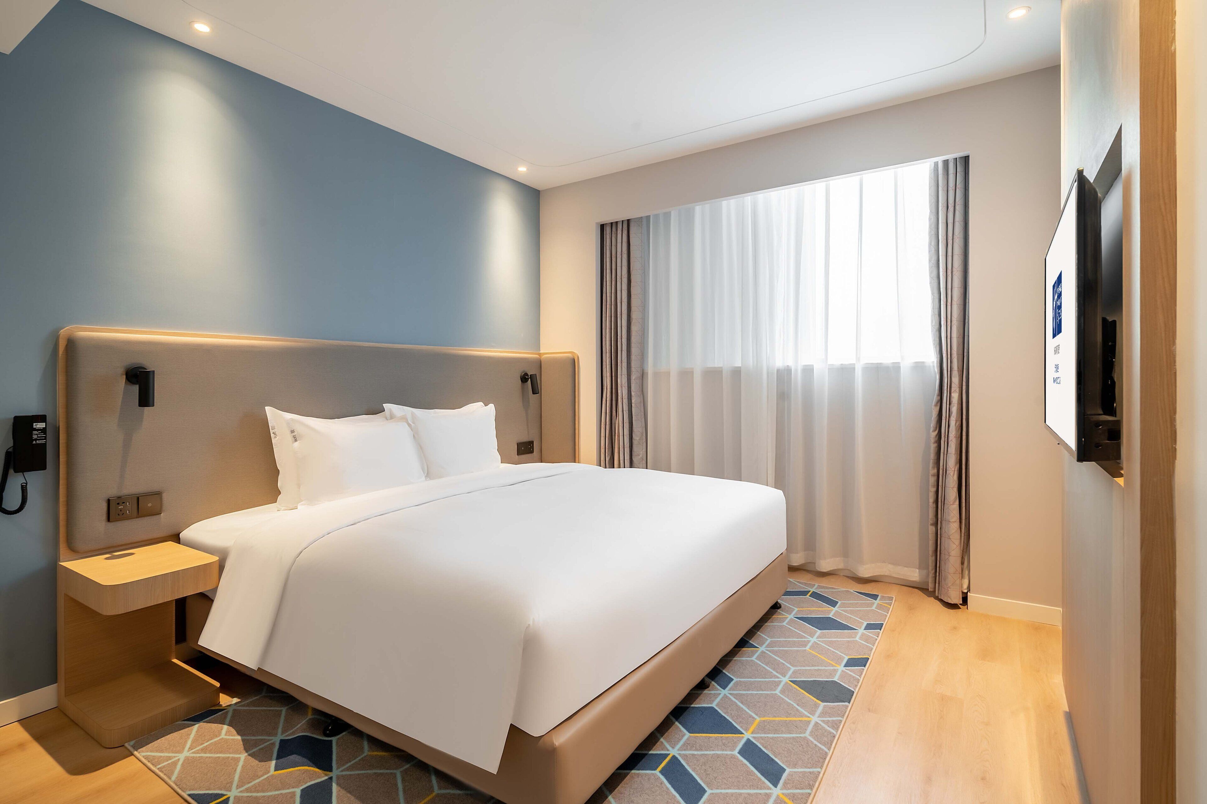 A modern hotel room with a large bed, a bedside table, a wall-mounted phone, and a window with curtains.