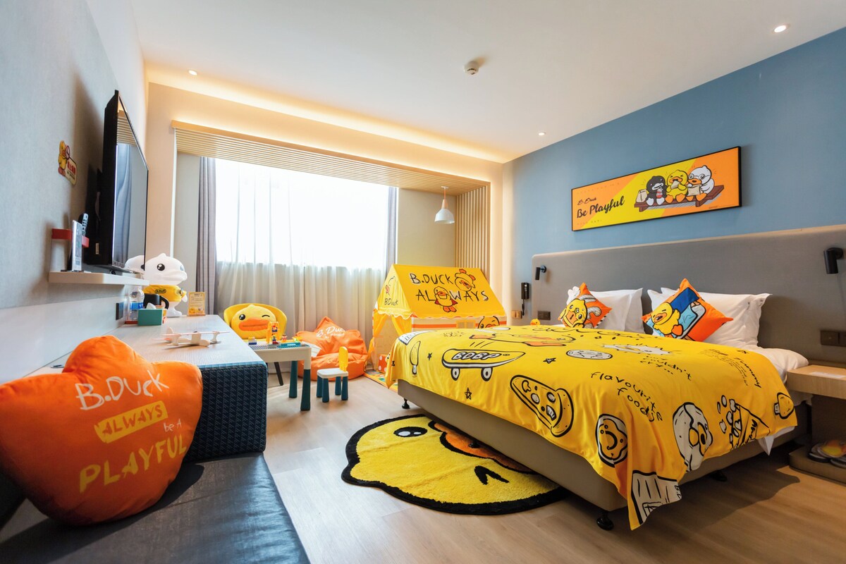 A hotel room with a bed covered in a yellow blanket with cartoon characters, a desk with a computer, a chair, and a window with curtains.